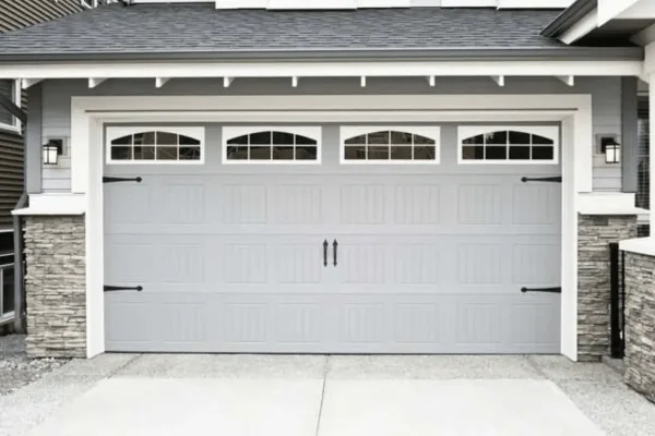 Wide gray garage door with four windows and wrought iron hinges and handles against a slate facade and asphalt shingle roof and concrete driveway