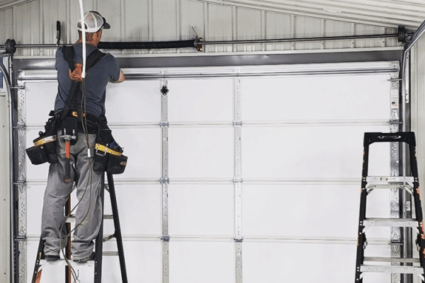 a garage door repairman adjusts the opening mechanism while standing on a stepladder in front of a white garage door